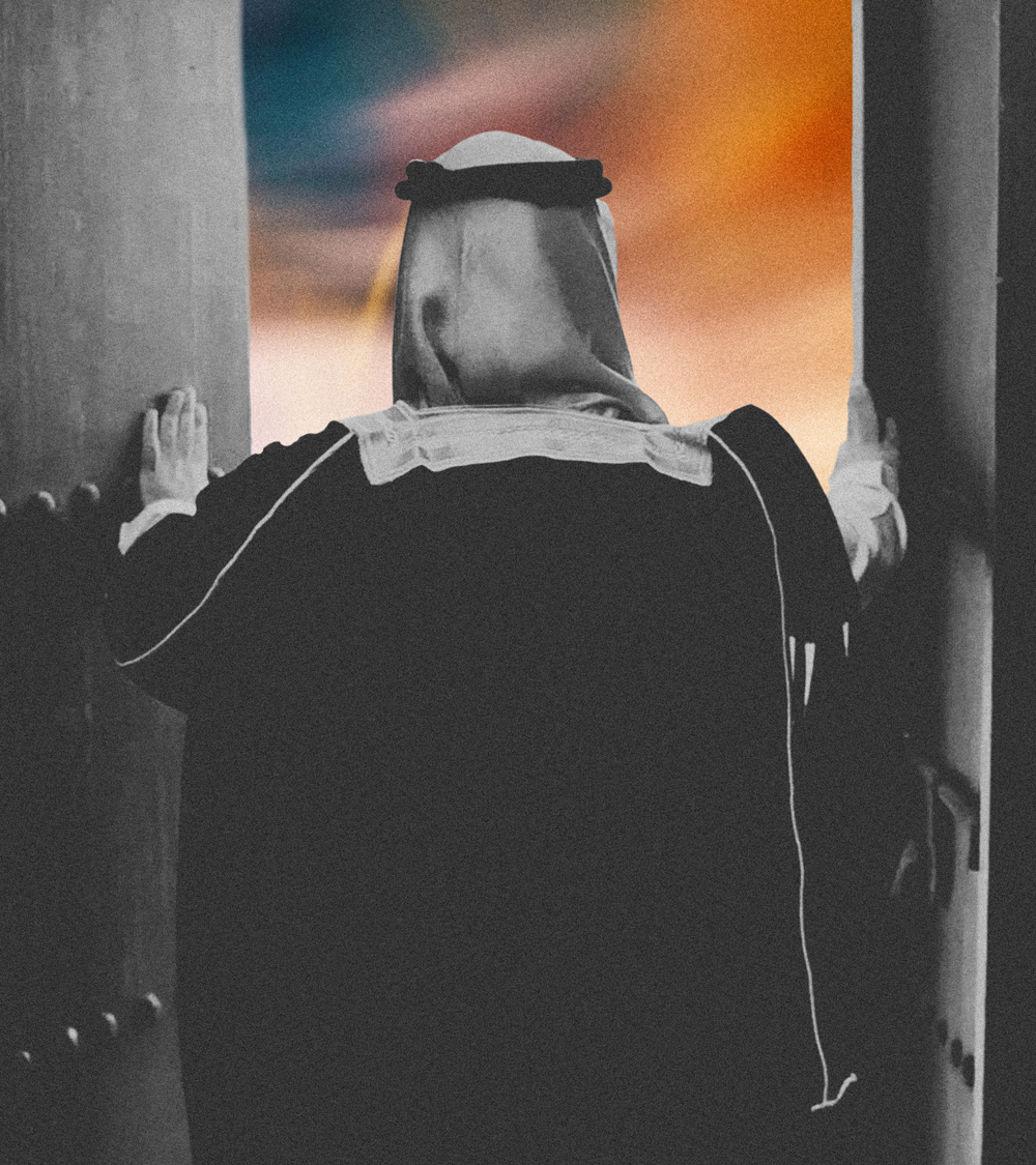 Man in traditional Saudi attire standing at a doorway, looking out toward a colorful sunset sky.